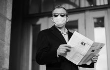 Caucasian man in sunglasses, medical mask and formal black suit, stands near building outdoor, holds newspaper in hands, poses at street, walks in quarantined city, finds out news from press